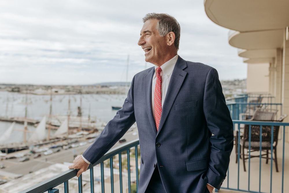 A groom in a dark suit and pink tie stands on an outdoor balcony overlooking a coastal landscape.