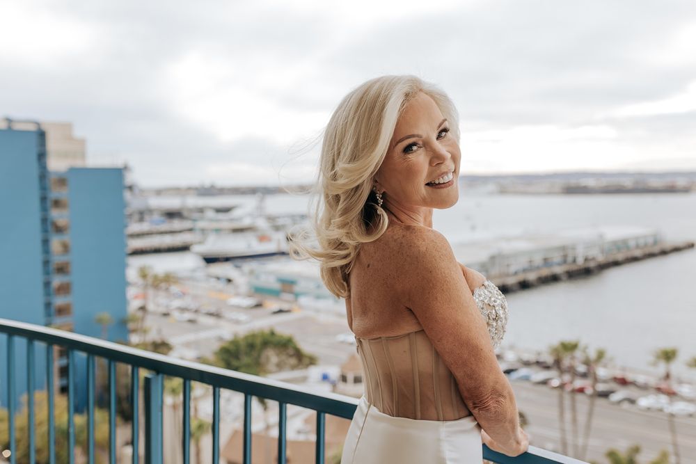 A bride with blonde hair stands on a balcony overlooking a marina with boats and water in the background.