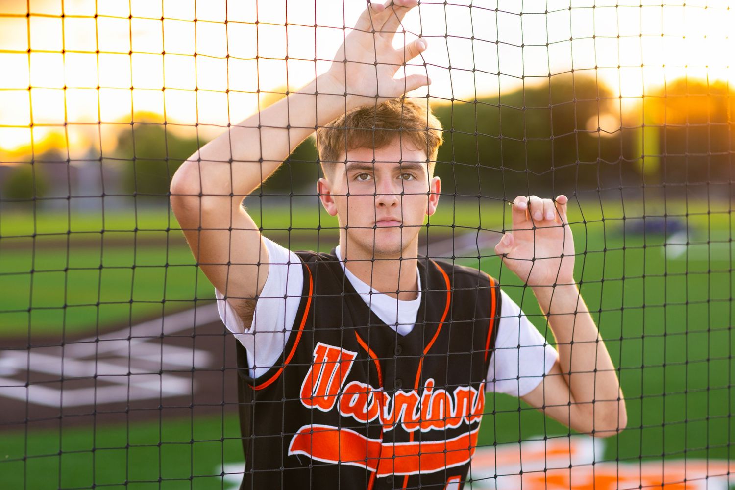 Senior Session of a Baseball player in black and orange uniform poses by batting cage netting at sunset.