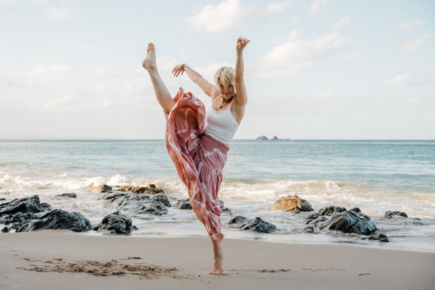 A dancer in a flowing pink skirt performs a graceful pose on a beach with volcanic rocks and ocean waves in the background.
