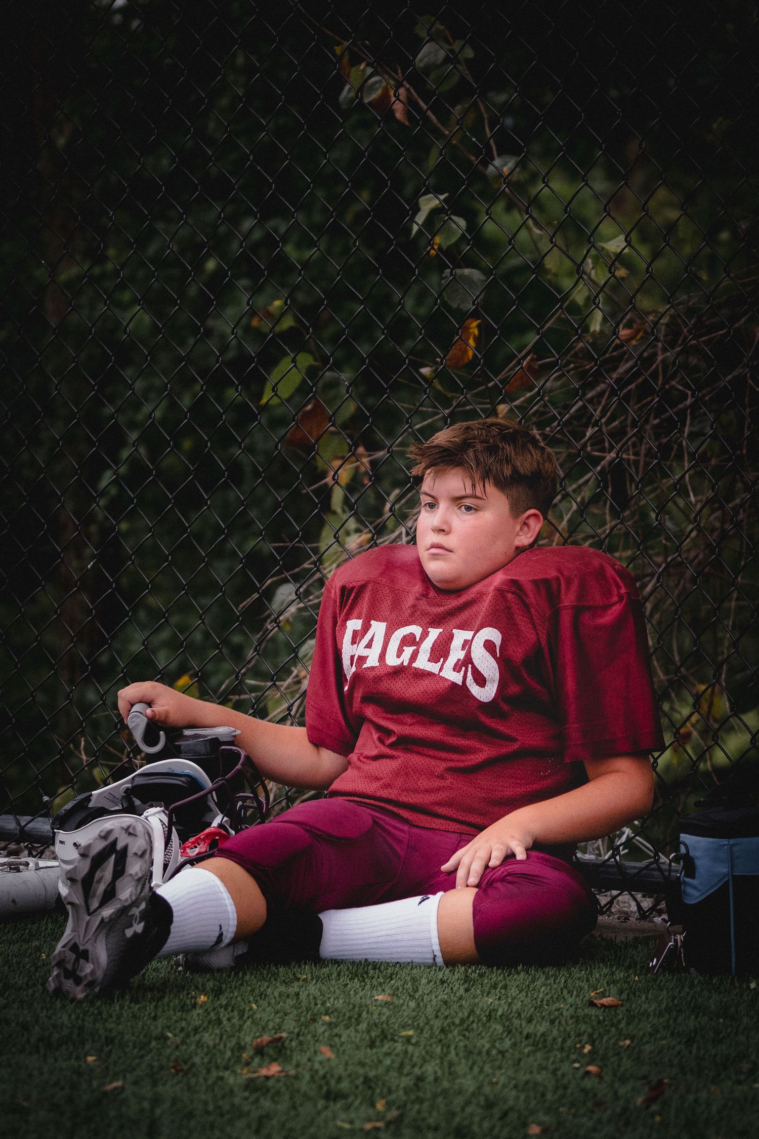 A young athlete in a maroon Eagles football uniform rests on field with sports equipment nearby.