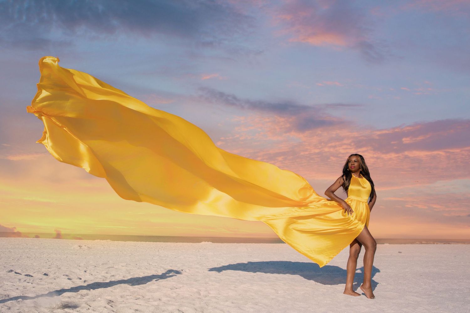 A figure in a flowing yellow dress poses dramatically against a sunset sky on a salt flat landscape.