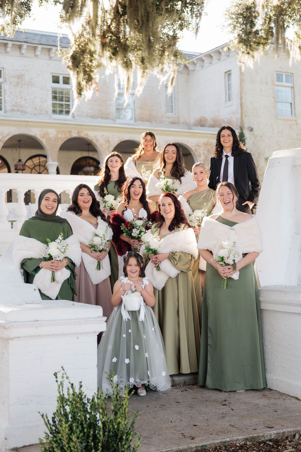 Wedding party in sage green dresses poses on white stone steps of an elegant Spanish-style building.