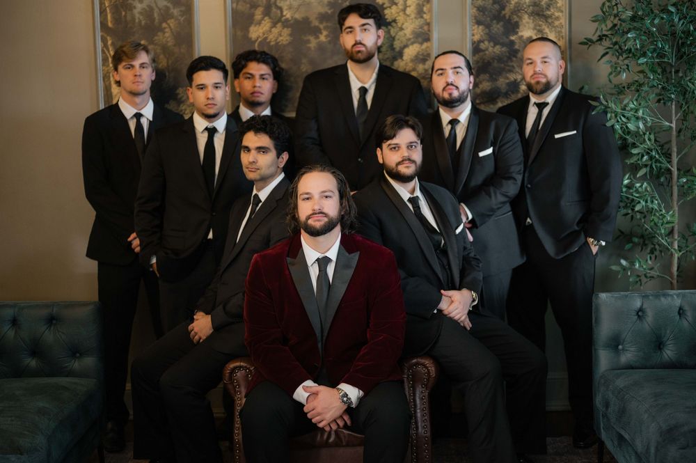 A group of groomsmen in formal suits and tuxedos pose together for a wedding party photo.