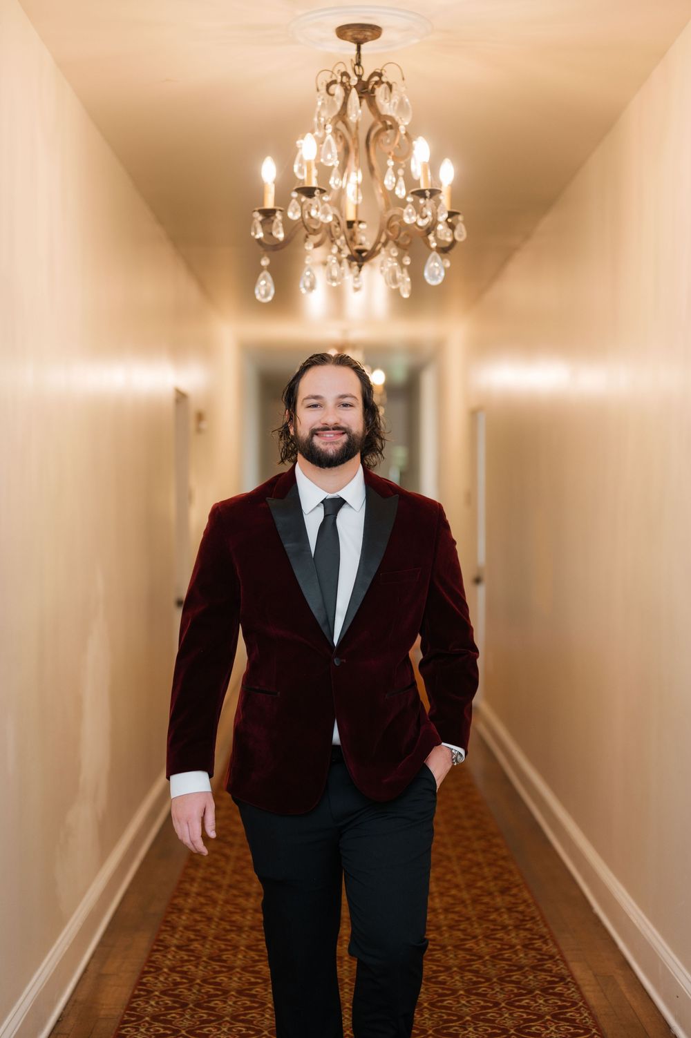A person in a dark suit walks down a carpeted hallway with chandeliers overhead.