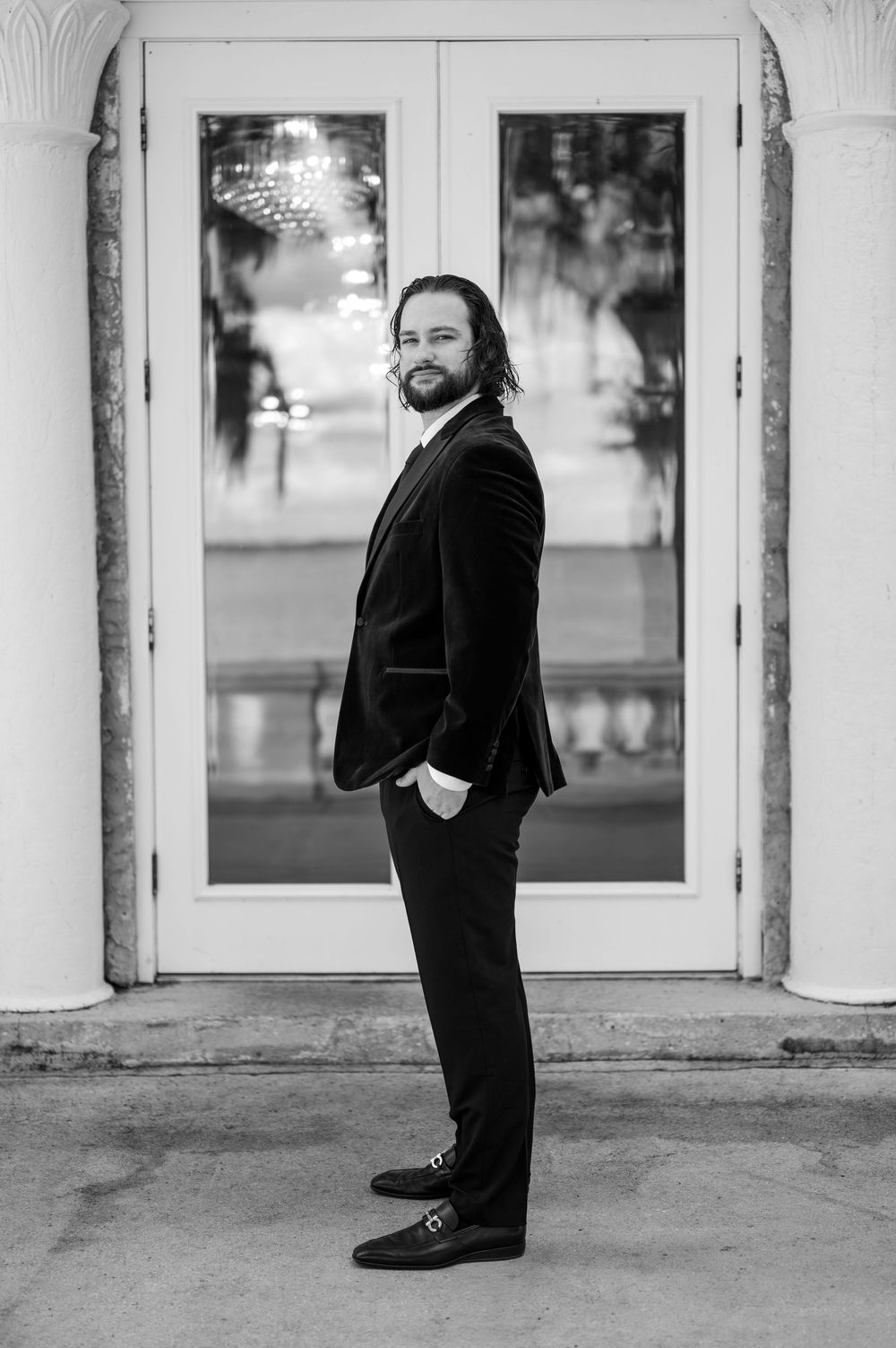 Black and white portrait of a bearded person in a formal suit standing in front of glass doors.