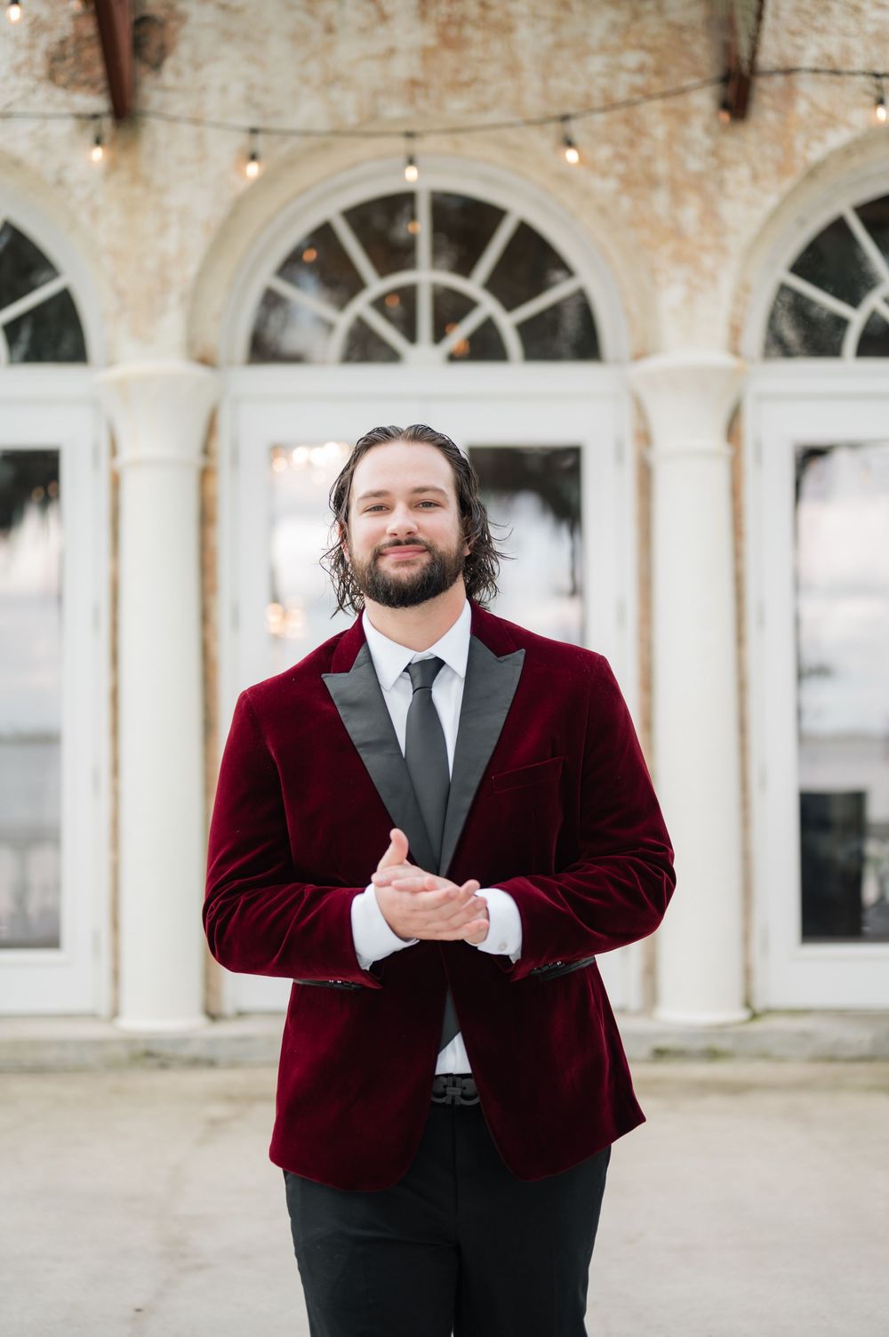 A groom in a burgundy velvet suit jacket poses emotionally against an ornate architectural backdrop.