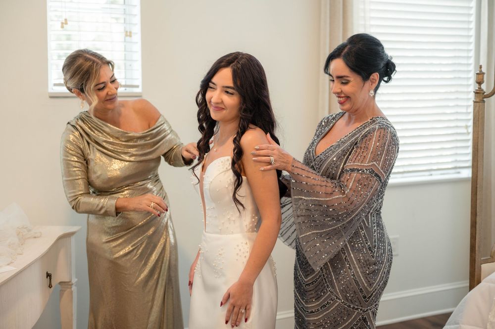 Three women in formal dresses help prepare for a special occasion in a sunlit room.