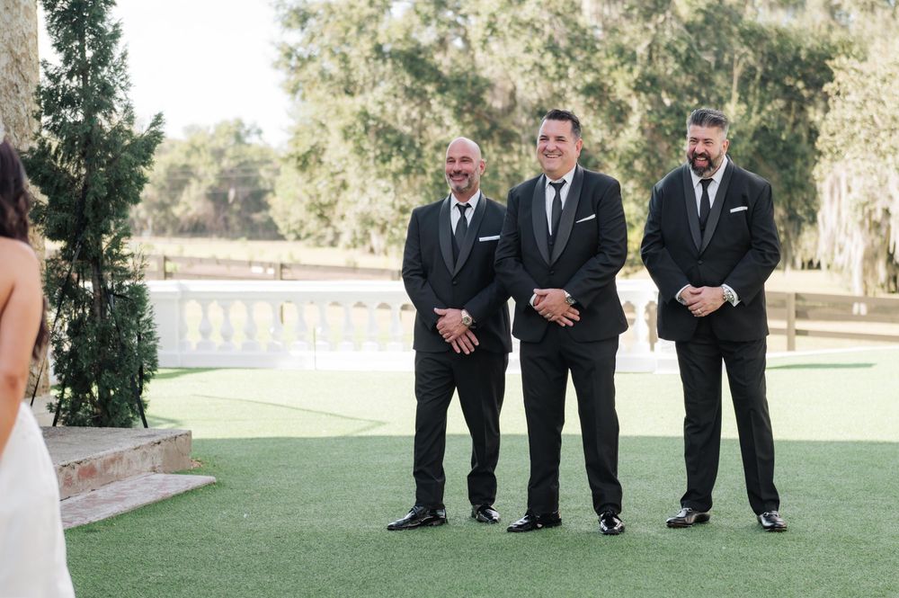 Three groomsmen in black suits stand on a manicured lawn during an outdoor wedding ceremony.