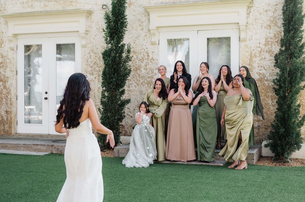 Wedding party in earth-toned dresses poses together on a grassy lawn against a stone building exterior.