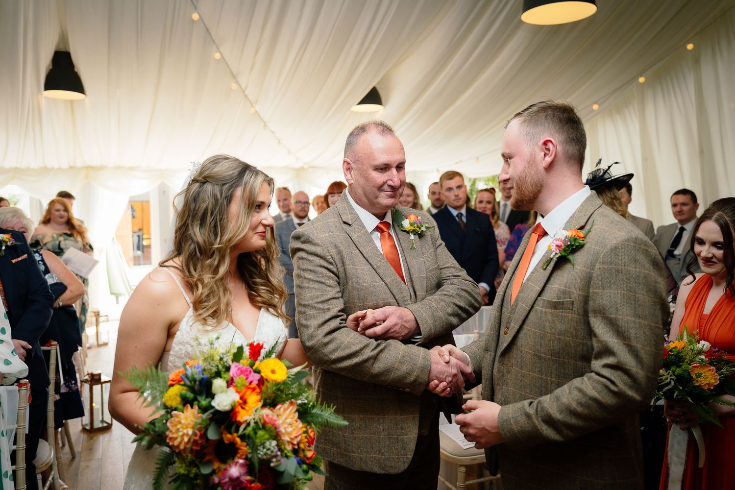 Emotional wedding ceremony moment with bright autumn bouquets inside white tent venue with hanging pendant lights.