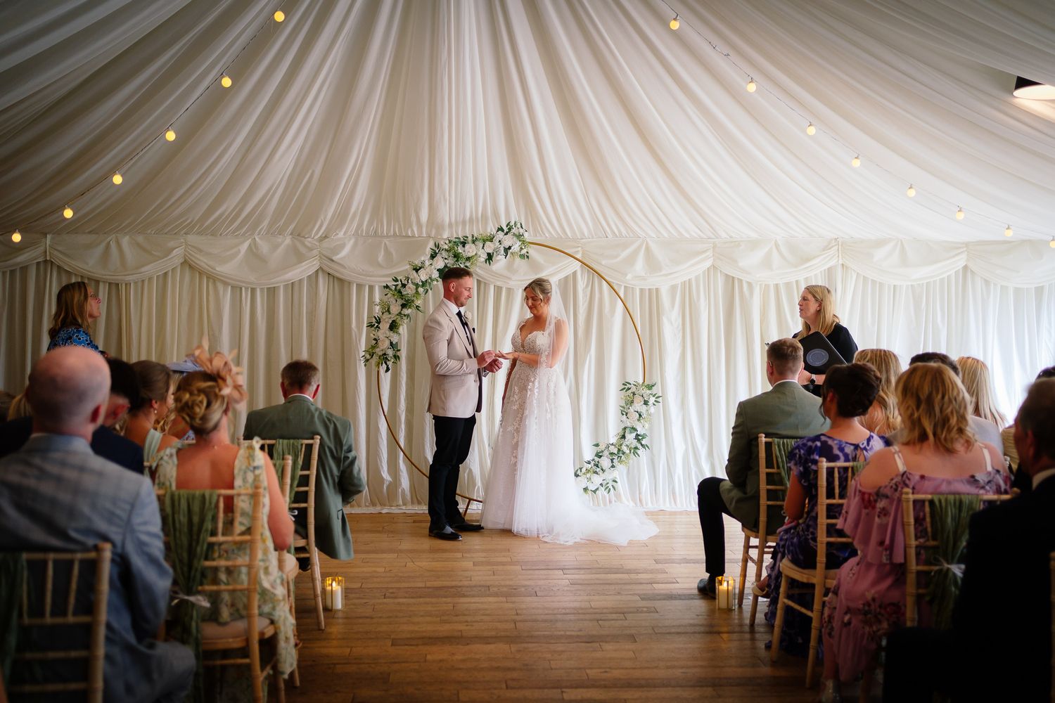 Wedding ceremony taking place inside an elegant white draped tent with guests seated in wooden chiavari chairs.