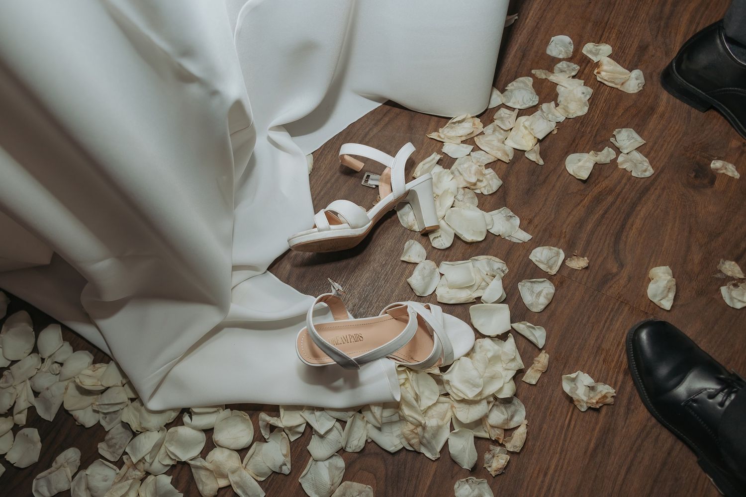 White rose petals scattered on hardwood floor next to a pair of white wedding heels and formal black shoes at San Clemente Shores.