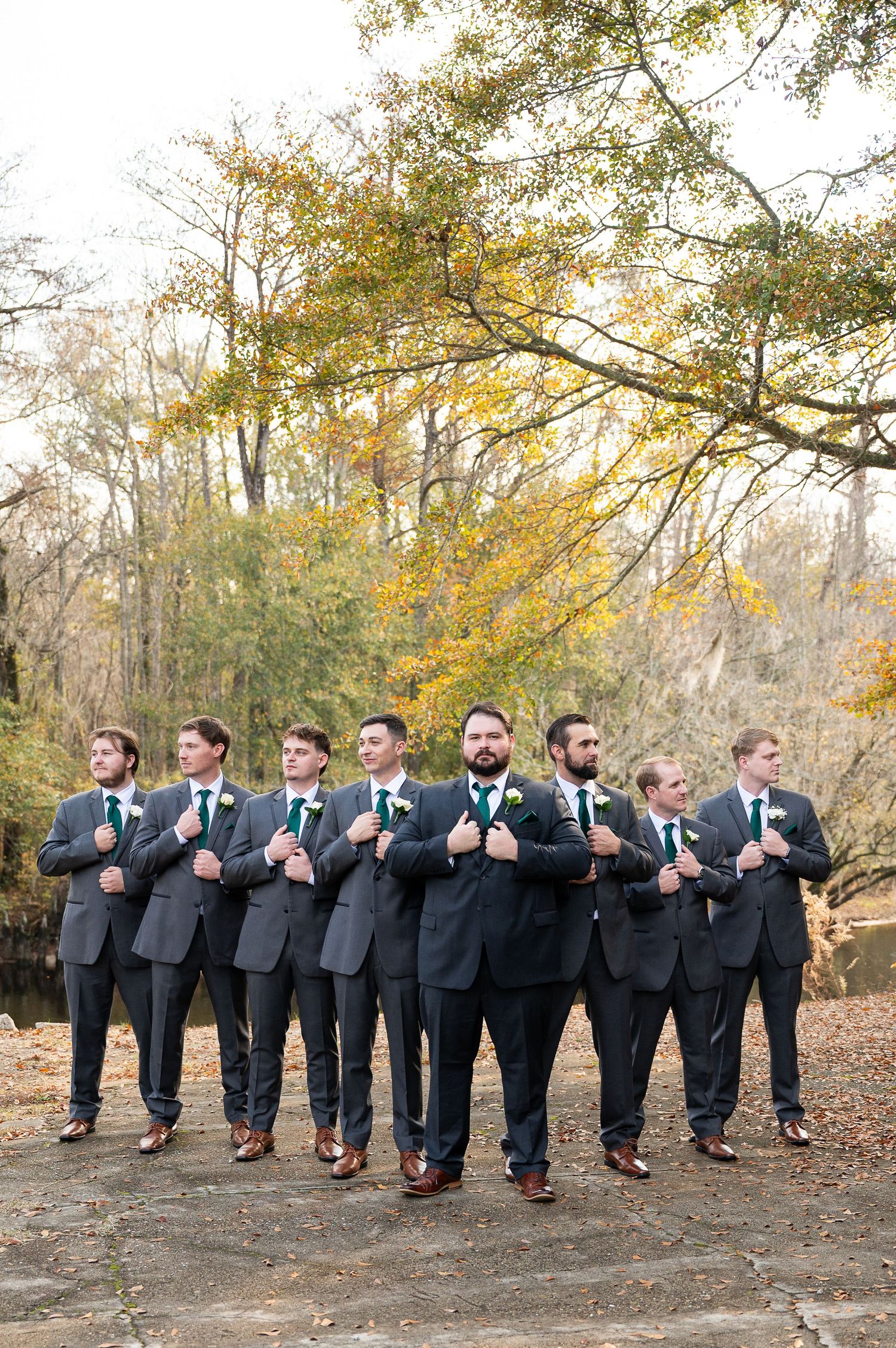 Groomsmen in dark suits pose in line with synchronized stance against autumn backdrop.