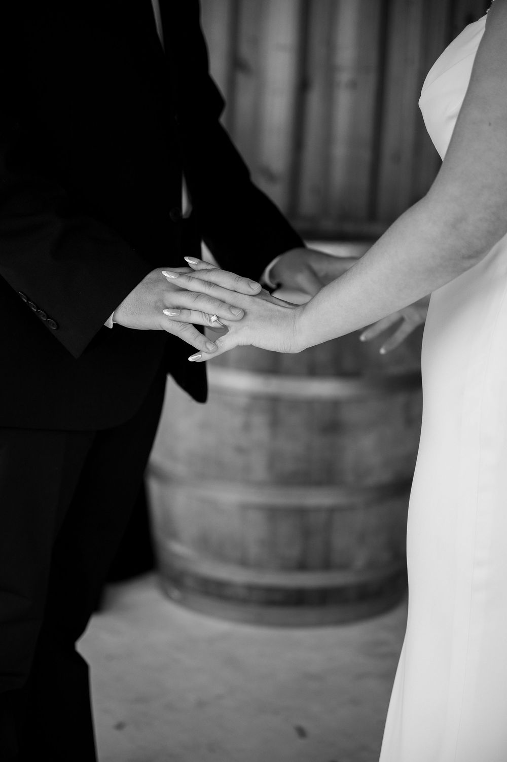 Black and white close-up of wedding rings being exchanged with a wooden barrel in background.