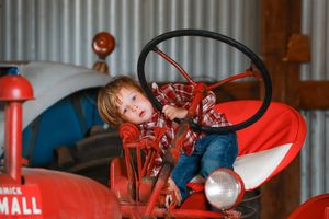 Child in red sweater sits playfully at the steering wheel of a vintage red tractor.