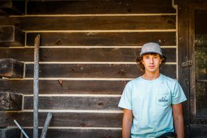 A person in a light blue t-shirt and baseball cap stands against a rustic wooden log cabin wall.