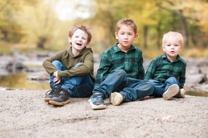 Three children in coordinating green outfits sit together on a sandy riverbank on an autumn day.