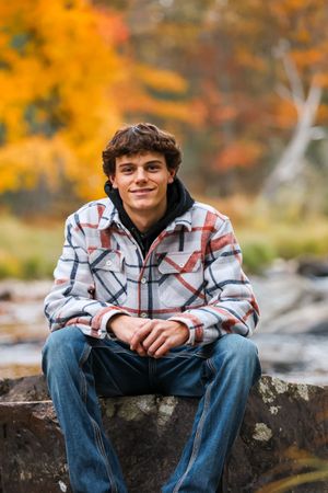 Person in plaid jacket sits on rocky ledge with vibrant autumn foliage creating warm orange backdrop.