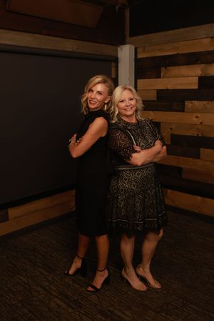 Two women in elegant evening wear pose playfully together in a rustic wood-paneled room.