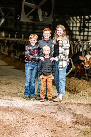 Three children stand together on a dirt path near a barn with farm animals visible in the background.