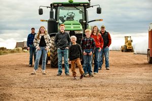 A group stands together on dirt ground in front of a large green John Deere tractor on a farm.