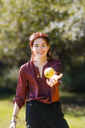 Someone wearing a burgundy top and floral crown holds a bright yellow lemon in a sunny outdoor setting.