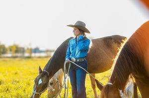 A person wearing a cowboy hat grooms a brown horse in a sunlit field with yellow wildflowers.