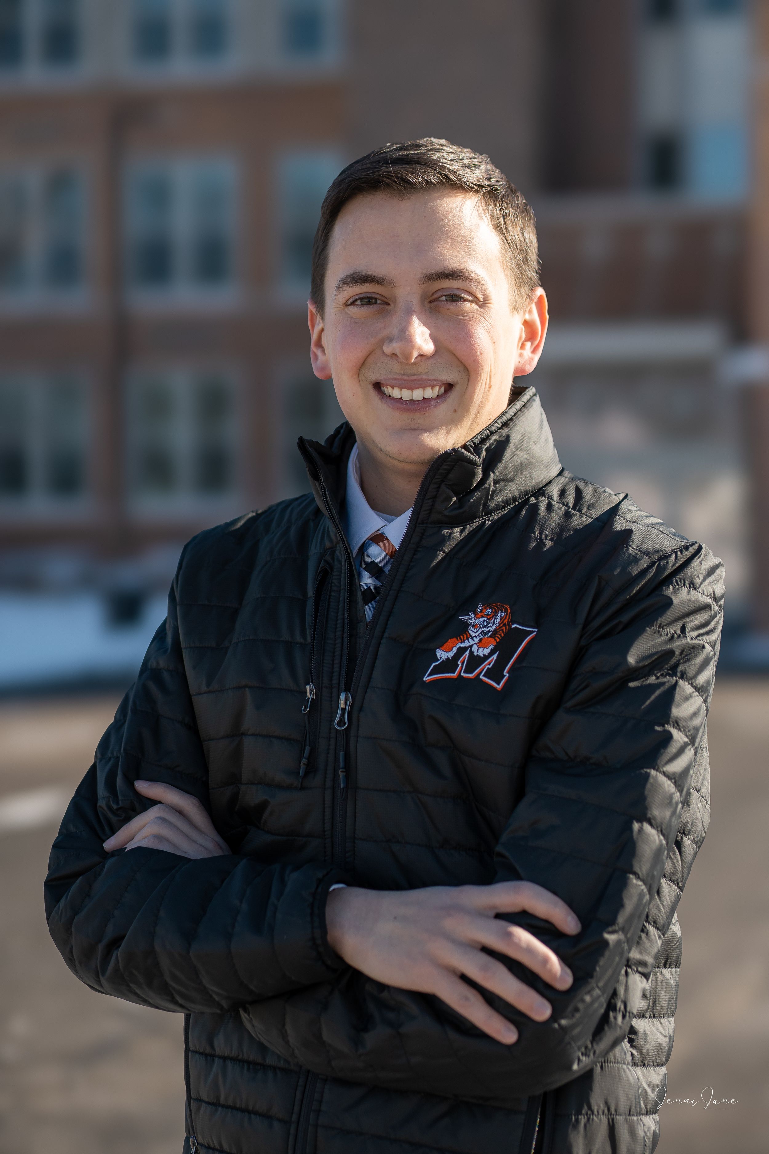 Professional outdoor portrait of someone wearing a black quilted jacket with a university logo and striped tie.