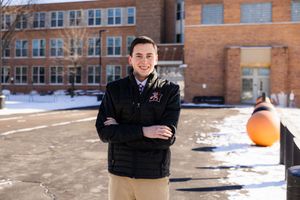 A person in winter attire stands in front of a brick school building with snow on the ground.