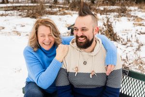 A couple sits together on a bench in a snowy winter landscape wearing matching cream sweaters and blue jackets.