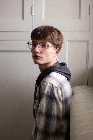 A person wearing glasses and a plaid shirt poses against a white wall in a series of moody portrait shots.