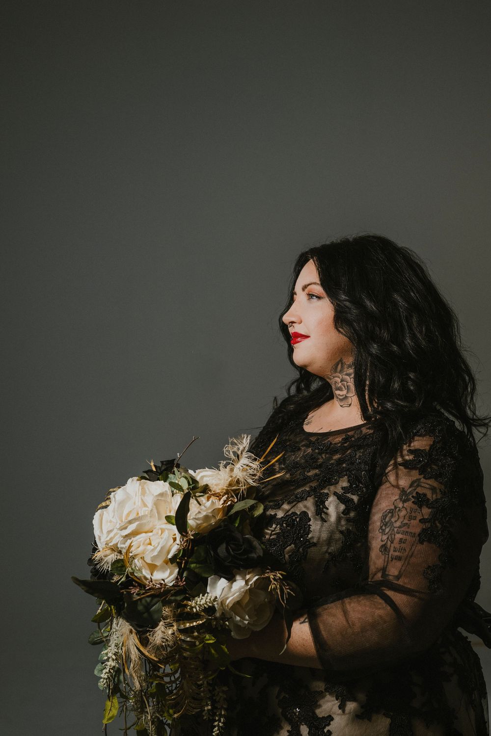 A dramatic portrait of a person in black lace holding a luxurious bouquet of cream-colored flowers against a dark background.