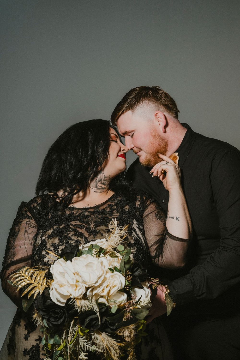 A romantic couple in black attire share intimate moments while holding an elegant white and gold floral bouquet.