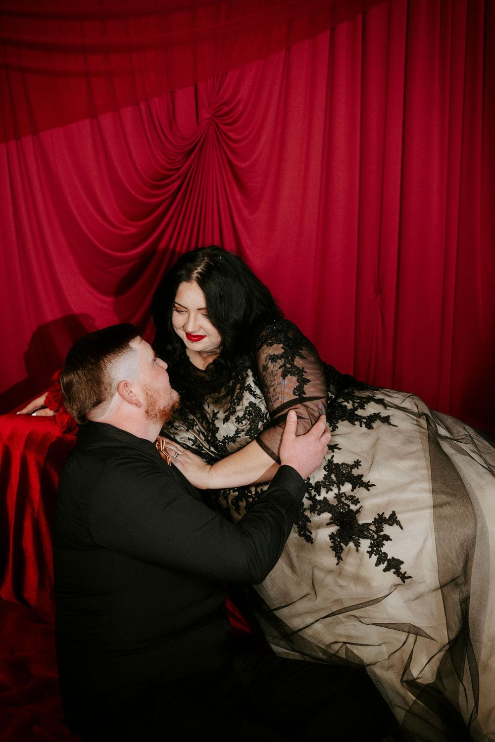 A romantic moment captured against red velvet curtains with a couple in formal attire embracing.