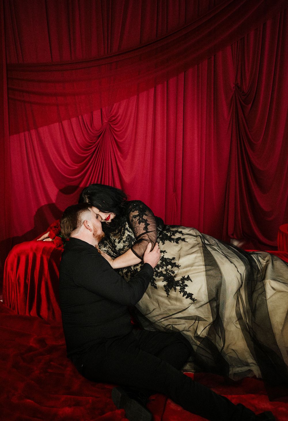 Figure in black reclines on beige chaise against red velvet curtains in vintage boudoir style photoshoot.