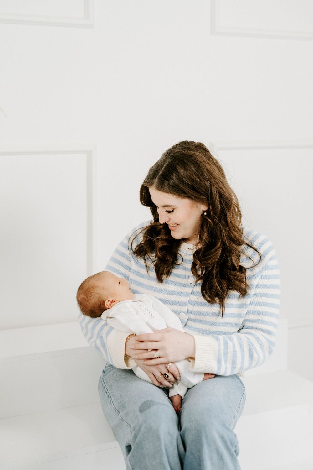 Parent in striped shirt holds and lovingly gazes at newborn baby in bright white minimalist setting.