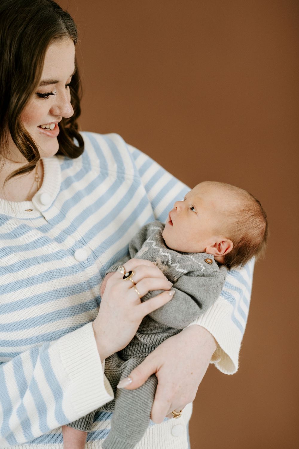 A tender moment between mother in blue striped sweater and young baby wearing grey outfit against brown backdrop.