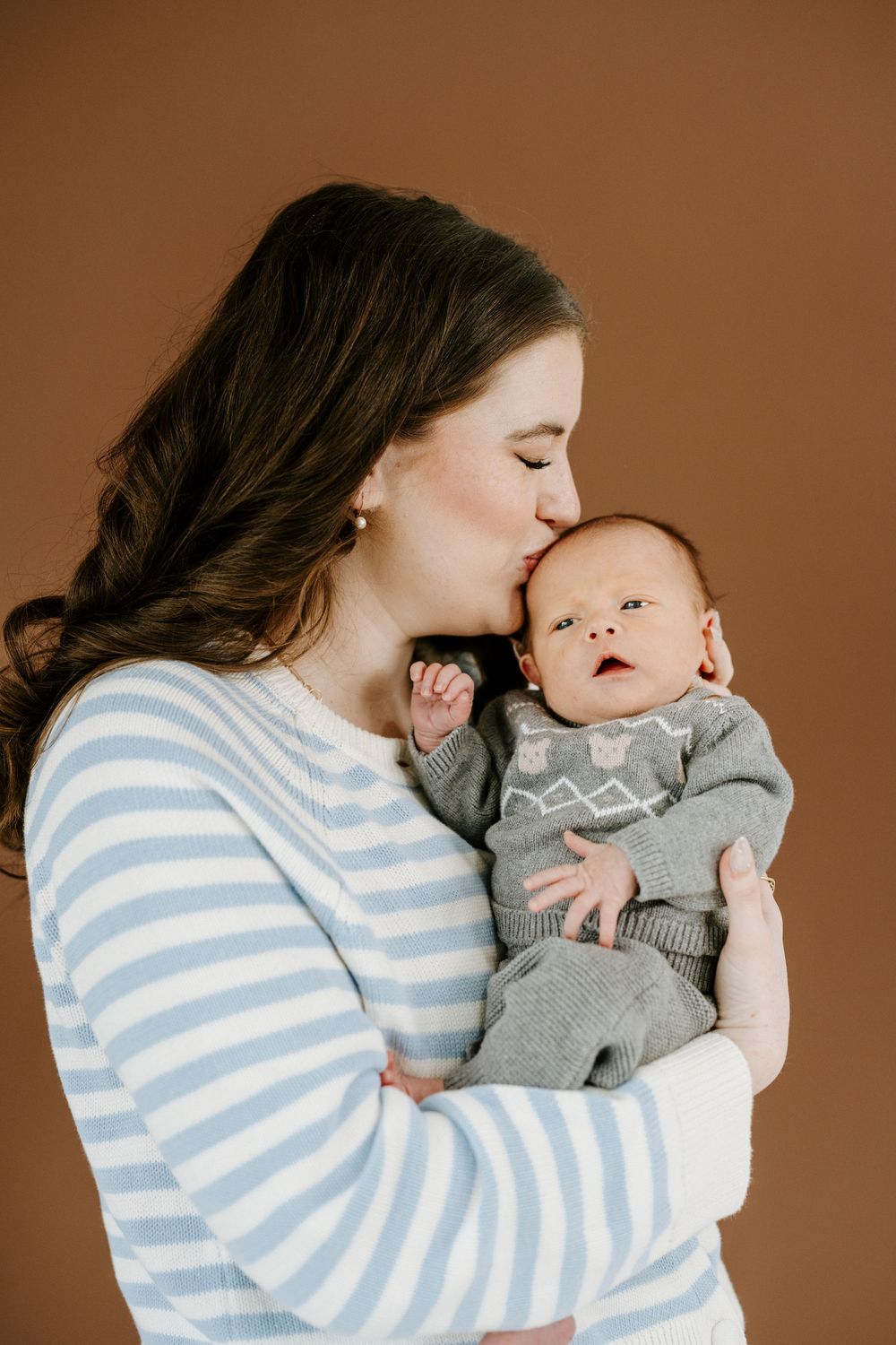 Intimate portrait of mother in striped sweater kissing her baby in grey sweater against brown background.