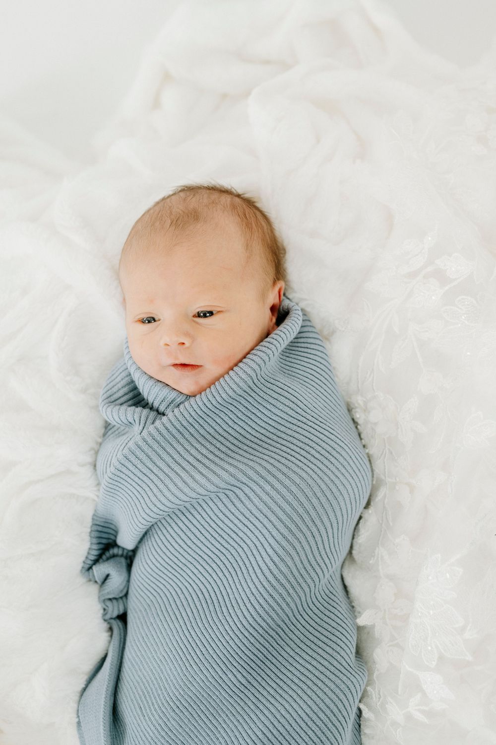 A tiny baby swaddled in a soft blue ribbed blanket lies on white fluffy fabric.