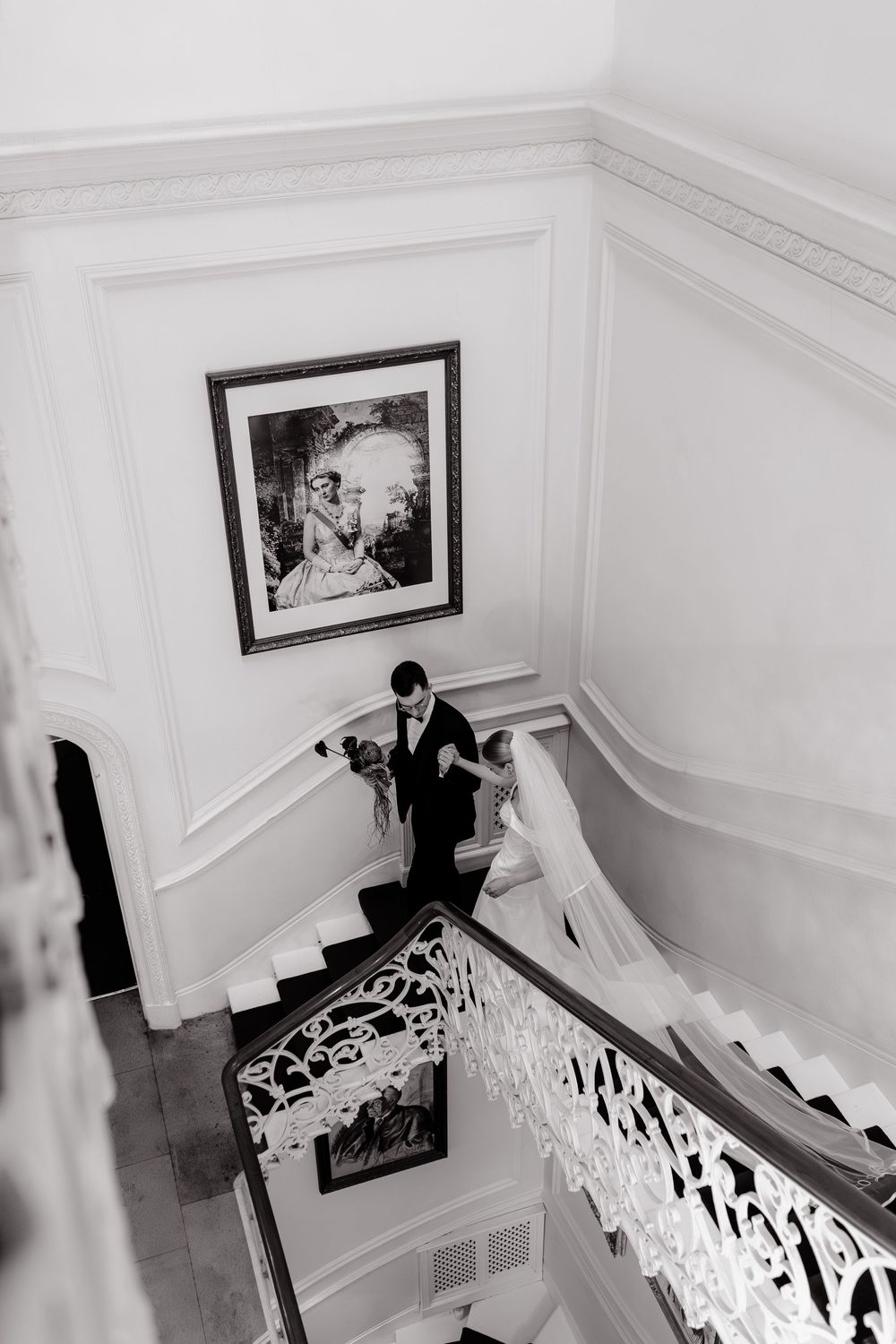 Black and white photograph showing a couple descending an elegant staircase with ornate white railings and framed artwork.
