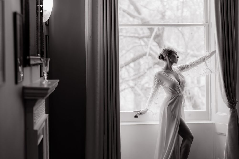 A bride in a flowing white dress stands near a window in a moody black and white photograph.