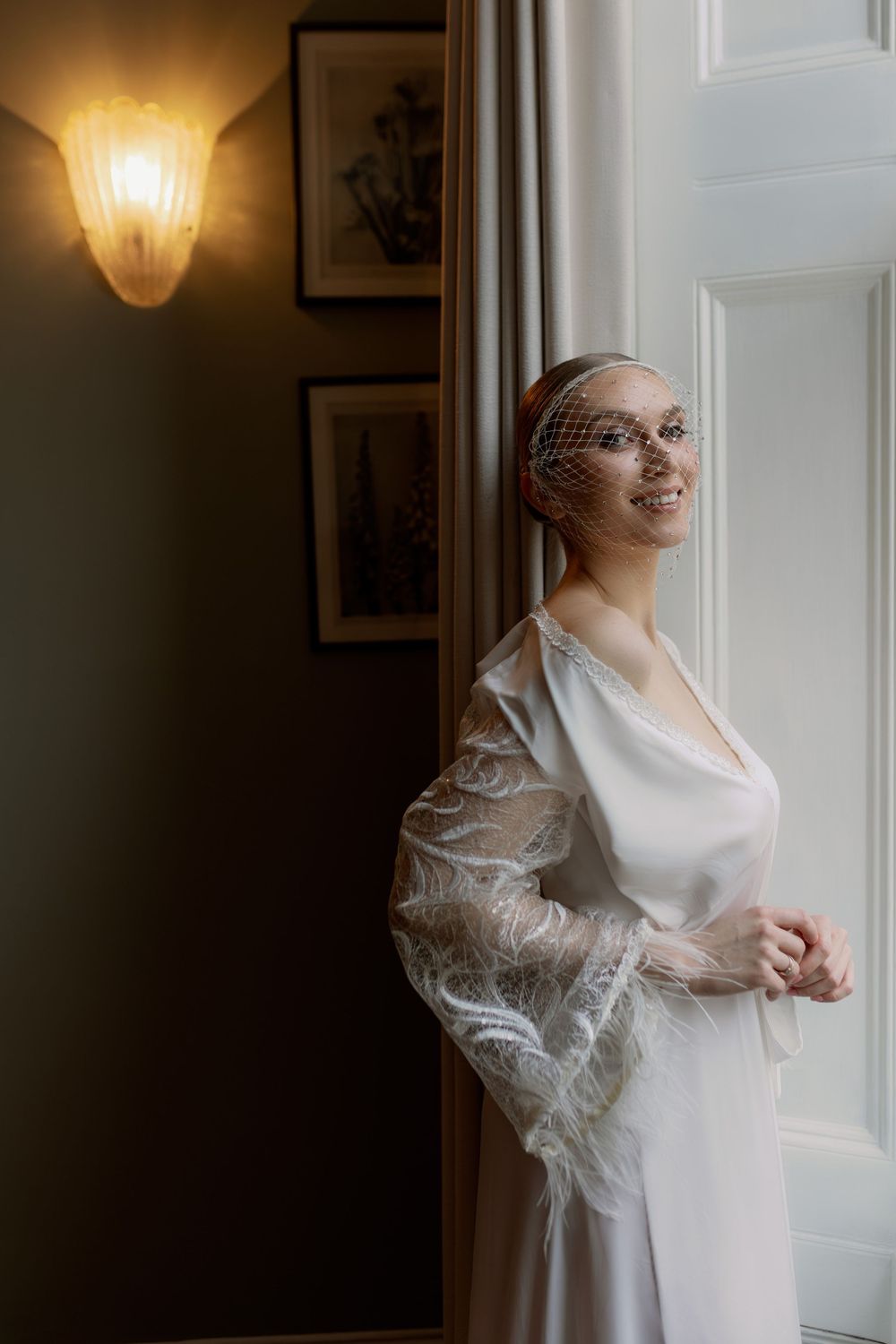 A bride in a vintage-style white gown with sheer sleeves stands by a window in soft lighting.