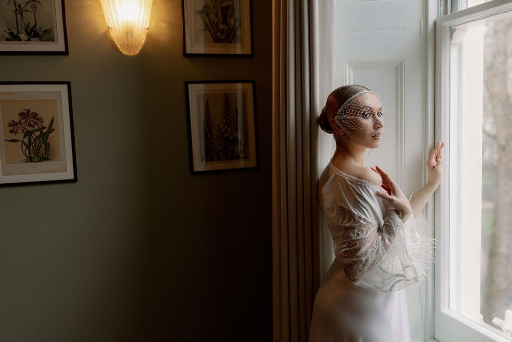 A person in a white dress stands contemplatively by a window in a room with framed artwork on the walls.