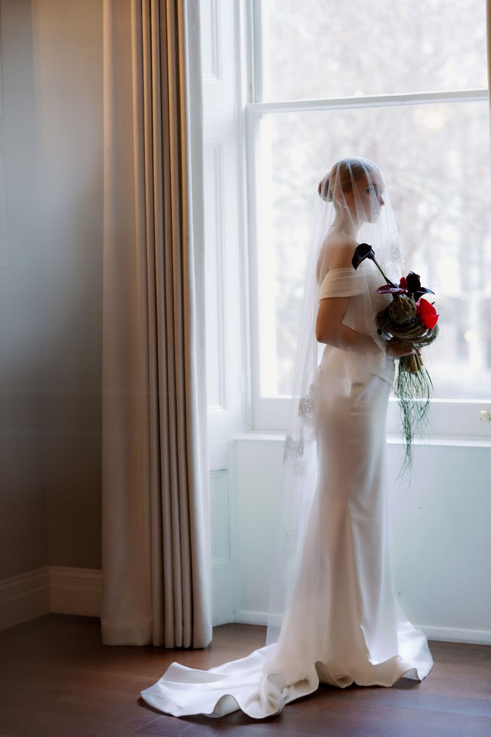 A silhouetted figure in a white wedding dress holds a red bouquet while standing by a bright window.