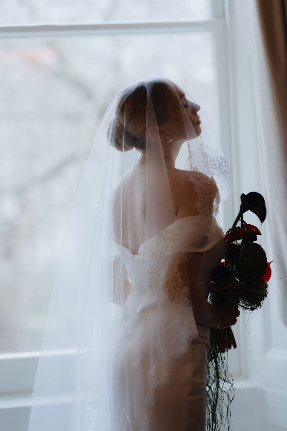 A silhouetted bride in a flowing white dress and veil stands gracefully by a window while holding a red rose bouquet.