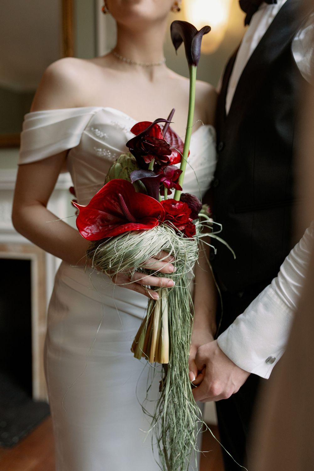 A bride in an off-shoulder white gown holds a striking bouquet of red calla lilies with cascading greenery.