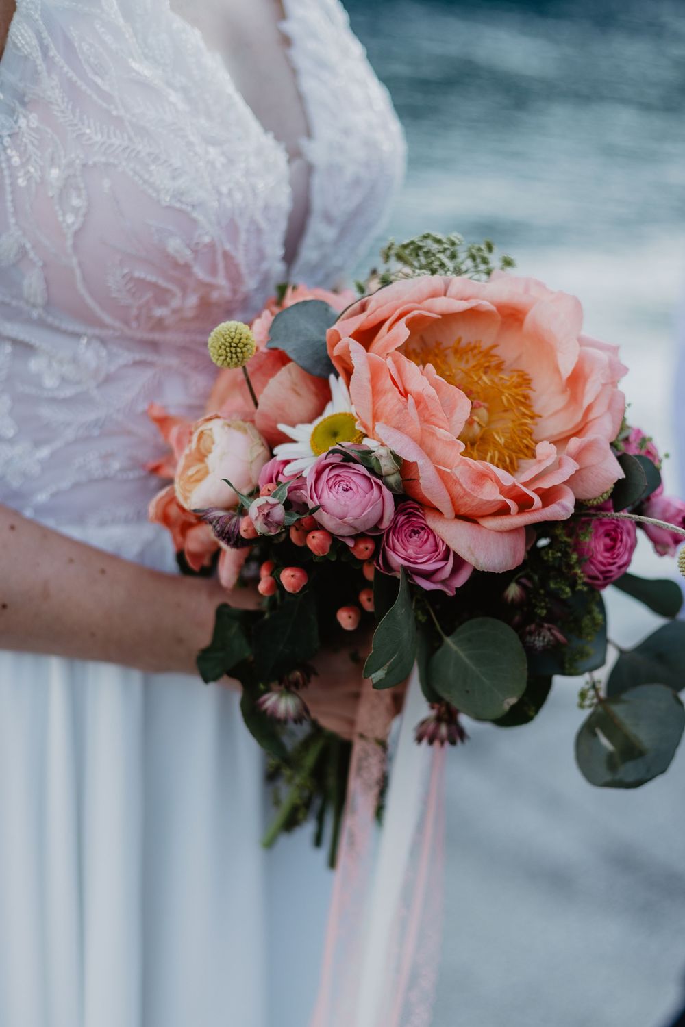 Delicate pink and coral floral bouquet with roses and greenery against a soft blue background.