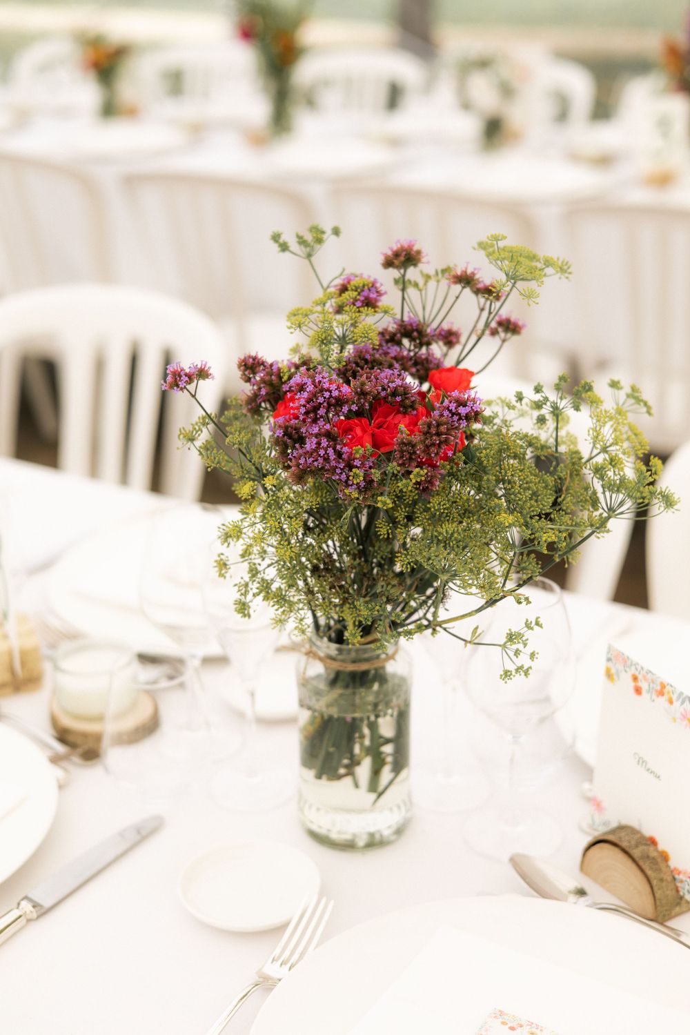 A rustic wedding centerpiece with wildflowers and red blooms in a mason jar on a white tablecloth.
