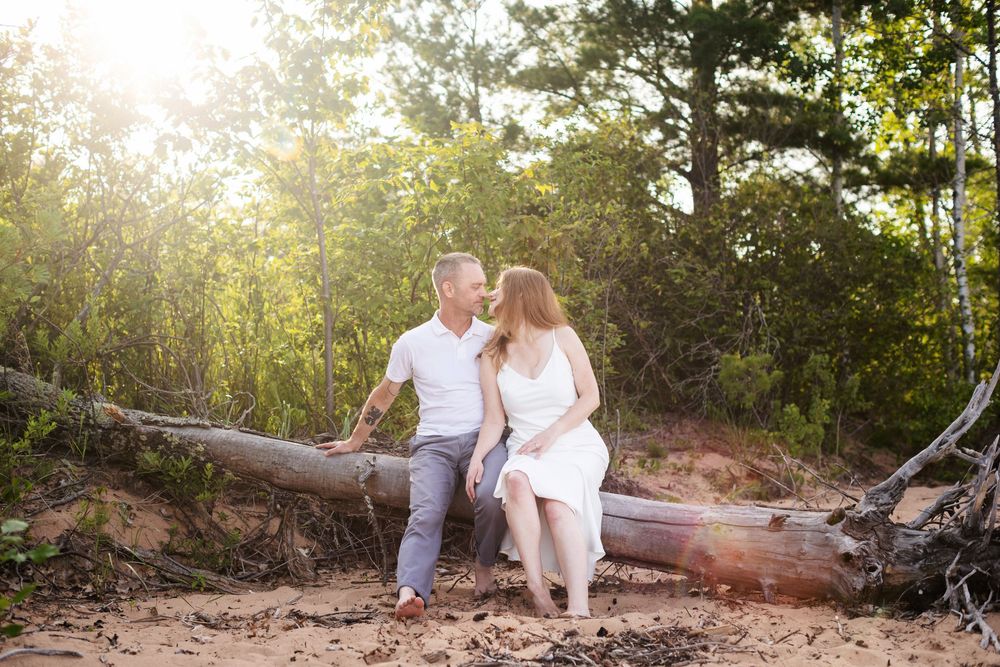 A couple sits together on a fallen log in a sunlit forest during a golden hour photoshoot.